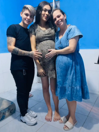 Uma grávida de vestido e pé descalço entre duas pessoas tocando em sua barriga